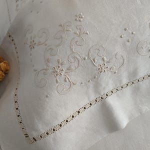 K&ouml;nnte beinhalten: Nahaufnahme eines wei&szlig;en Kopfkissenbezugs mit aufwendigen Stickereien in hellbeige. Das Design umfasst florale und wirbelnde Muster. Der Kissenbezug hat eine dekorative Bord&uuml;re mit kleinen, gleichm&auml;&szlig;ig verteilten Ausschnitten, die einen eleganten Look erzeugt.