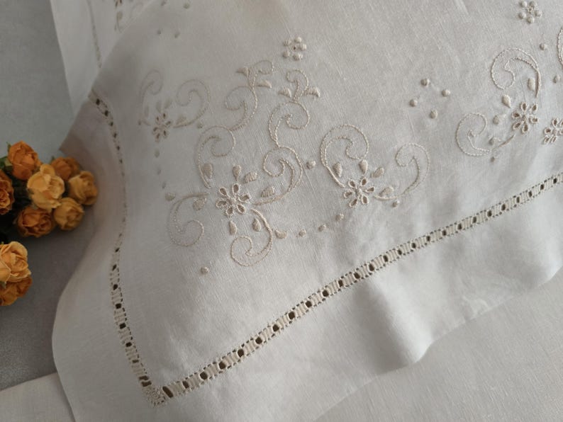 K&ouml;nnte beinhalten: Nahaufnahme eines wei&szlig;en Kopfkissenbezugs mit aufwendigen Stickereien in hellbeige. Das Design umfasst florale und wirbelnde Muster. Der Kissenbezug hat eine dekorative Bord&uuml;re mit kleinen, gleichm&auml;&szlig;ig verteilten Ausschnitten, die einen eleganten Look erzeugt.
