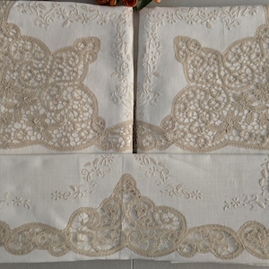 May include: White linen bedding set with intricate beige lace detailing. The set includes pillowcases and a sheet, all adorned with delicate floral embroidery and scalloped edges. The lace design adds a touch of elegance to the bedroom decor.