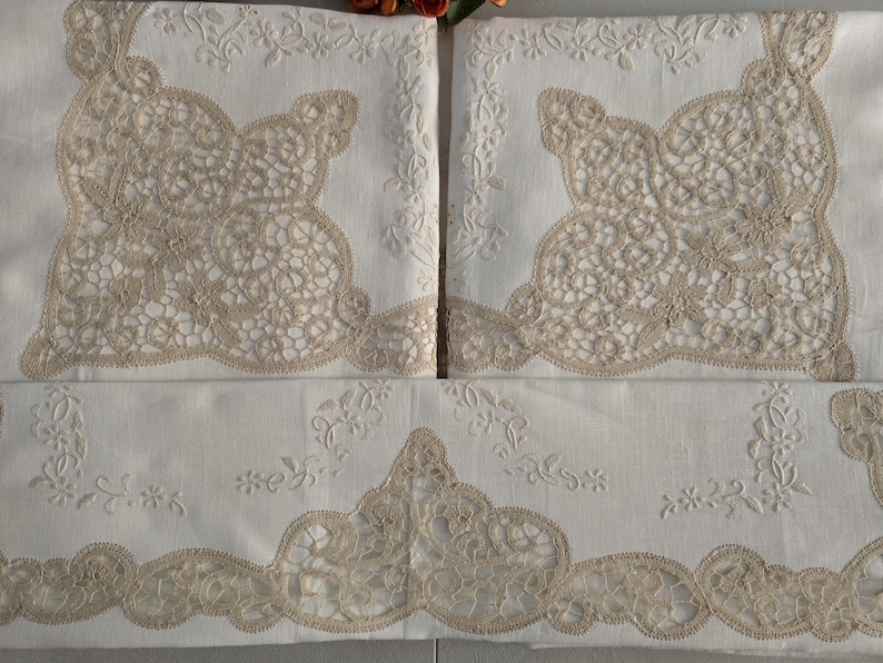 May include: White linen bedding set with intricate beige lace detailing. The set includes pillowcases and a sheet, all adorned with delicate floral embroidery and scalloped edges. The lace design adds a touch of elegance to the bedroom decor.
