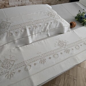 May include: White embroidered bedding set, including a pillowcase and a long sheet, with detailed floral and geometric patterns. The bedding is arranged on a bed, with a small bouquet of blue flowers and a decorative ball.