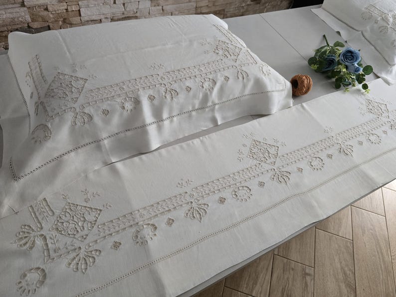 May include: White embroidered bedding set, including a pillowcase and a long sheet, with detailed floral and geometric patterns. The bedding is arranged on a bed, with a small bouquet of blue flowers and a decorative ball.