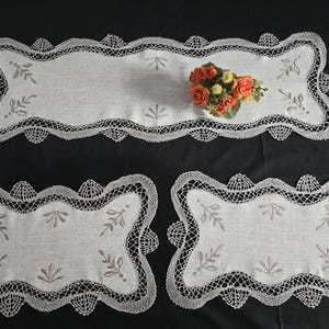 Puede incluir: Tres manteles de encaje blanco con bordados delicados. Los manteles tienen bordes festoneados y presentan un diseño floral.
