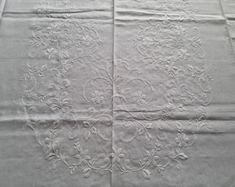 May include: White linen tablecloth with an embroidered floral design. The design features delicate white flowers and leaves arranged in a symmetrical pattern.