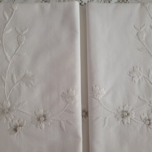 May include: Two white pillowcases with delicate embroidered floral designs. The embroidery features white flowers and vines on a white background.