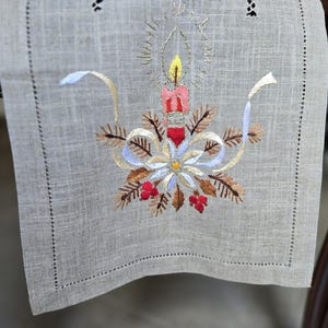 Pode incluir: Caminho de mesa de linho bordado com um desenho de vela. A vela tem uma chama amarela, um corpo vermelho e rosa e detalhes decorativos. O desenho inclui folhas castanhas, fitas brancas e azuis e bagas vermelhas. O caminho de mesa é cinzento claro.