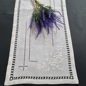 May include: White linen table runner with delicate embroidered floral designs and a scalloped edge. The runner is decorated with a floral pattern and has a white background.