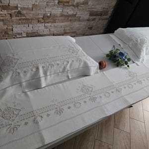 May include: White embroidered bedding set with pillowcases, a sheet, and a runner. The set features detailed floral and geometric patterns. A small bouquet of blue flowers and a decorative item are placed on the bed.