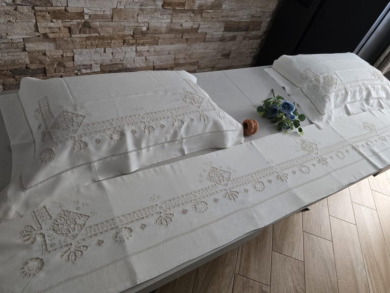 May include: White embroidered bedding set with pillowcases, a sheet, and a runner. The set features detailed floral and geometric patterns. A small bouquet of blue flowers and a decorative item are placed on the bed.