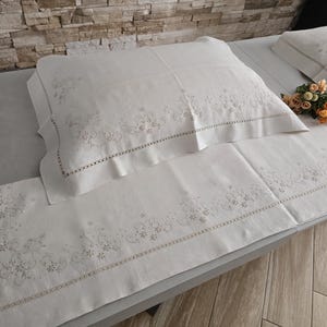 K&ouml;nnte beinhalten: Wei&szlig;es besticktes Bettw&auml;sche-Set mit floralen Mustern. Das Set umfasst ein Kissen und ein langes Laken, beide mit zarten Stickereien an den R&auml;ndern. Ein kleiner Blumenstrau&szlig; liegt auf dem Bett. Die Bettw&auml;sche ist aus weichem Material.