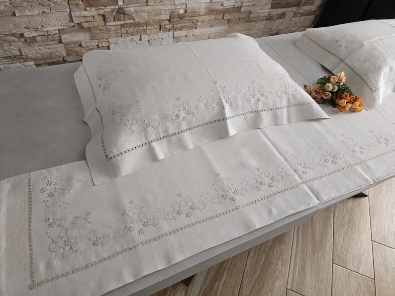 K&ouml;nnte beinhalten: Wei&szlig;es besticktes Bettw&auml;sche-Set mit floralen Mustern. Das Set umfasst ein Kissen und ein langes Laken, beide mit zarten Stickereien an den R&auml;ndern. Ein kleiner Blumenstrau&szlig; liegt auf dem Bett. Die Bettw&auml;sche ist aus weichem Material.