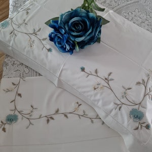 May include: White bed linen set featuring embroidered floral patterns in blue, beige, and grey. A large blue rose adorns the pillowcase. The set includes a pillowcase and a sheet, offering a delicate and elegant appearance.