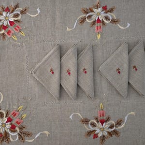 Puede incluir: Un mantel de lino beige y servilletas a juego con diseños bordados de flores y velas navideñas. Las servilletas están dobladas en triángulos y presentan un pequeño diseño bordado de bayas rojas y ramitas. El mantel tiene detalles bordados de flores y cintas.
