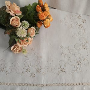 K&ouml;nnte beinhalten: Wei&szlig;es Leinentischtuch mit gesticktem Blumen- und Rankenmuster. Ein Strau&szlig; aus pfirsichfarbenen und orangenen Rosen und Gr&uuml;n liegt auf der Ecke. Das Tischtuch hat einen dekorativen Rand.