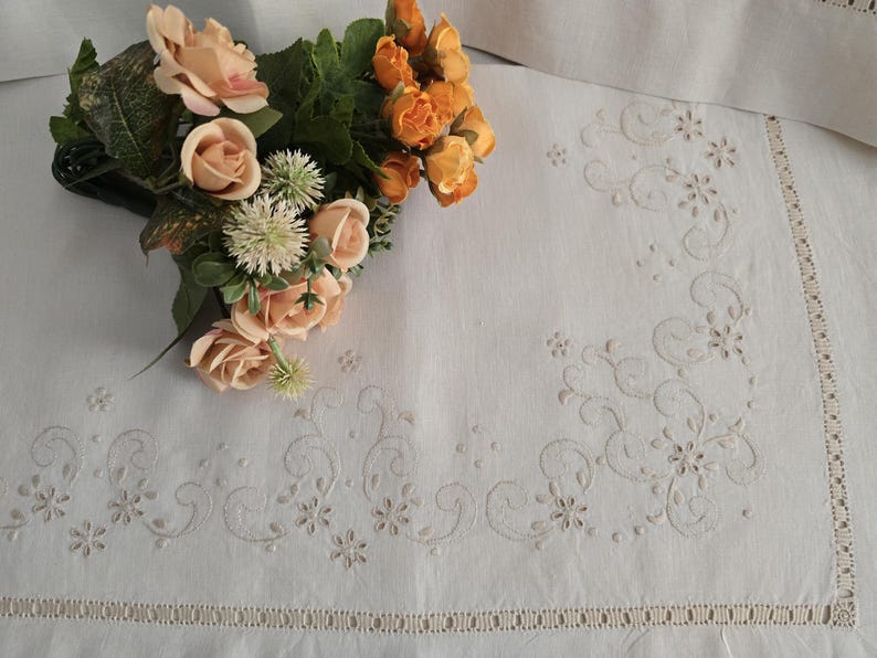 K&ouml;nnte beinhalten: Wei&szlig;es Leinentischtuch mit gesticktem Blumen- und Rankenmuster. Ein Strau&szlig; aus pfirsichfarbenen und orangenen Rosen und Gr&uuml;n liegt auf der Ecke. Das Tischtuch hat einen dekorativen Rand.