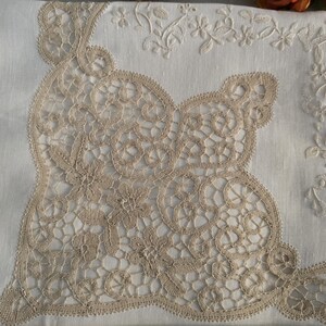 May include: Close-up of a white linen textile with intricate beige lace detailing. The lace forms a large, ornate design in the center, with floral embroidery along the edges. The overall aesthetic is delicate and vintage.