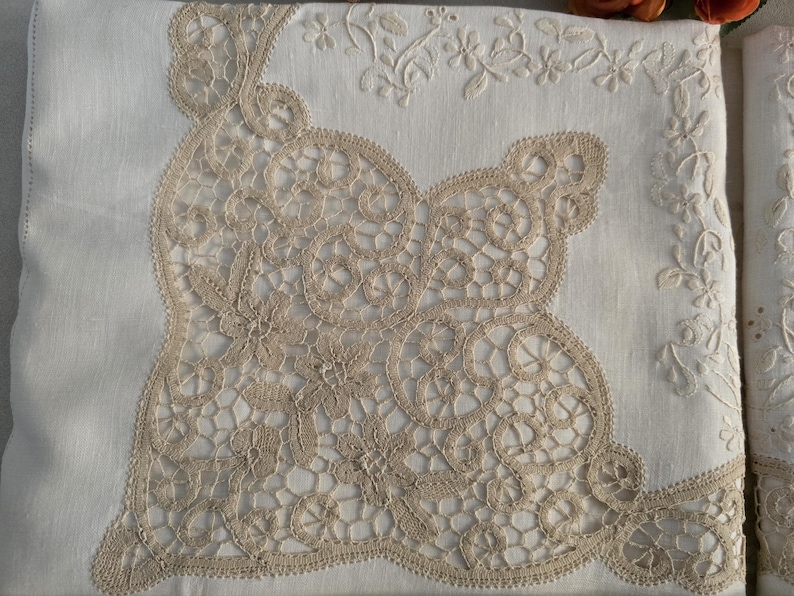 May include: Close-up of a white linen textile with intricate beige lace detailing. The lace forms a large, ornate design in the center, with floral embroidery along the edges. The overall aesthetic is delicate and vintage.