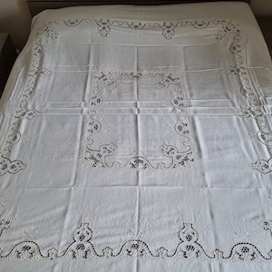 Può includere: Tovaglia bianca con ricami traforati intricati. Il design presenta un motivo ripetuto di forme floreali e geometriche, con uno sfondo blu scuro visibile attraverso le aperture. La tovaglia è stesa su un letto.