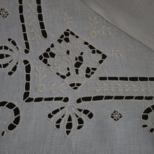 May include: Close-up of a white linen tablecloth featuring detailed cutwork and embroidered floral and geometric patterns. The cutwork creates open spaces, and the embroidery showcases delicate floral designs. The tablecloth has a classic, elegant aesthetic.
