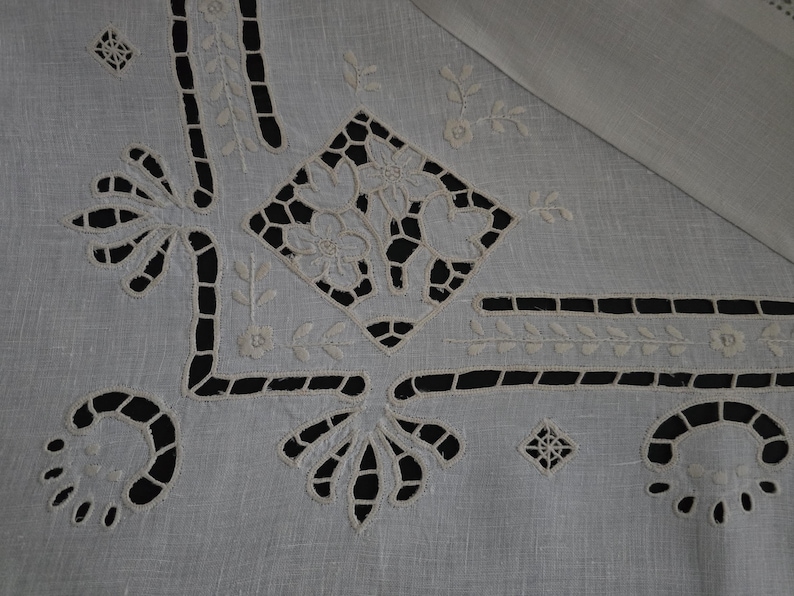 May include: Close-up of a white linen tablecloth featuring detailed cutwork and embroidered floral and geometric patterns. The cutwork creates open spaces, and the embroidery showcases delicate floral designs. The tablecloth has a classic, elegant aesthetic.