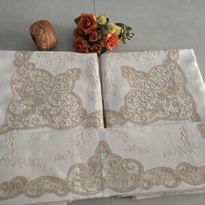 May include: White linen tablecloths with intricate beige lace detailing. The tablecloths are folded and arranged, showcasing the delicate floral and lace patterns. A small bouquet of orange roses and a ball of twine are also present.