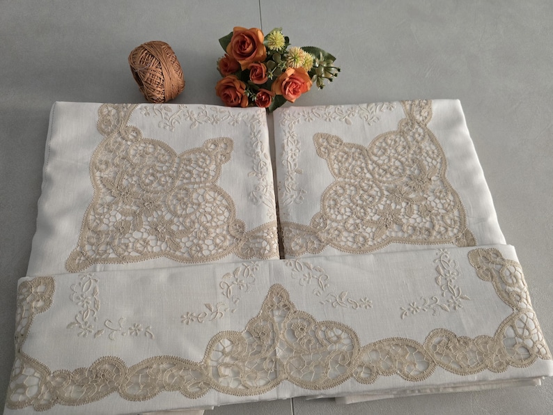 May include: White linen tablecloths with intricate beige lace detailing. The tablecloths are folded and arranged, showcasing the delicate floral and lace patterns. A small bouquet of orange roses and a ball of twine are also present.