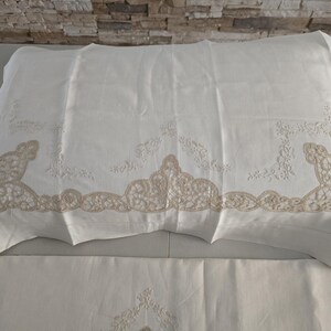 May include: White pillowcase with beige lace trim and floral embroidery. The pillowcase is made of a soft, textured fabric and is folded over a white sheet. The lace trim features a detailed, ornate design.