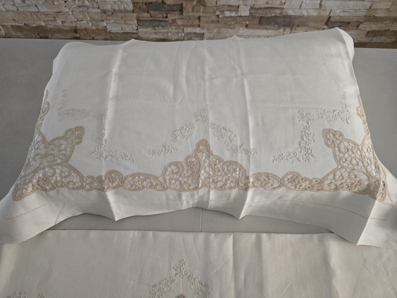 May include: White pillowcase with beige lace trim and floral embroidery. The pillowcase is made of a soft, textured fabric and is folded over a white sheet. The lace trim features a detailed, ornate design.