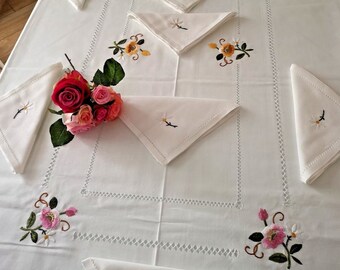 May include: White embroidered tablecloth with a delicate floral design. The tablecloth features pink and yellow flowers with green leaves. The tablecloth is set with white napkins, also embroidered with floral designs.