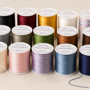 May include: A collection of fifteen spools of 100% silk pearl stringing thread in various colors including black, burgundy, gold, white, grey, olive, brown, cream, pink, lavender, blue, and navy, each labeled with the text 100% Silk Pearl Stringing Size: F.