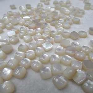 May include: A large group of white, iridescent, square-shaped, mother of pearl cabochons. The cabochons are scattered on a white surface.