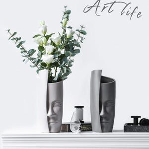 May include: Two face-shaped vases, one white and one gray, sit on a white shelf. The white vase holds white flowers and green foliage. The gray vase is empty. The words "Art life" are written in cursive above the vases.