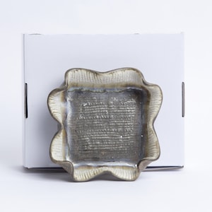 以下が含まれることがあります： 波打つ縁のある小さな正方形のセラミック皿。皿は暗いグレーで、微妙な虹色の光沢のある釉薬がかかっています。