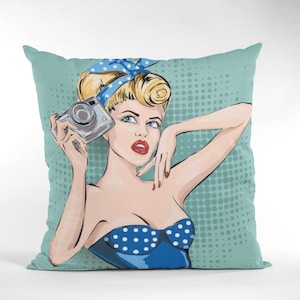 Peut inclure: Un coussin de décoration bleu sarcelle avec une illustration pop art d'une femme aux cheveux blonds et une robe bleue à pois blancs tenant un appareil photo. L'arrière-plan est bleu sarcelle avec des pois blancs.