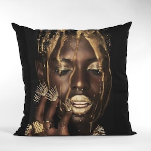 May include: A black and gold throw pillow with a close-up image of a person's face covered in gold paint.