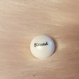 Puede incluir: Piedra redonda blanca con la palabra "Blessed" escrita en negro.