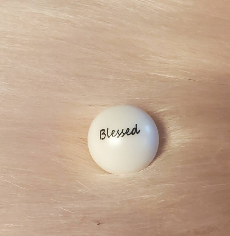Puede incluir: Piedra redonda blanca con la palabra "Blessed" escrita en negro.
