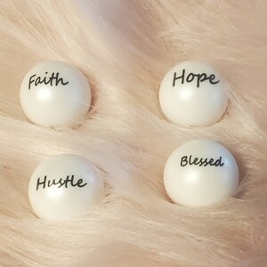 Puede incluir: Cuatro adornos redondos blancos con letras negras. Las palabras en los adornos son "Faith", "Hope", "Hustle" y "Blessed".