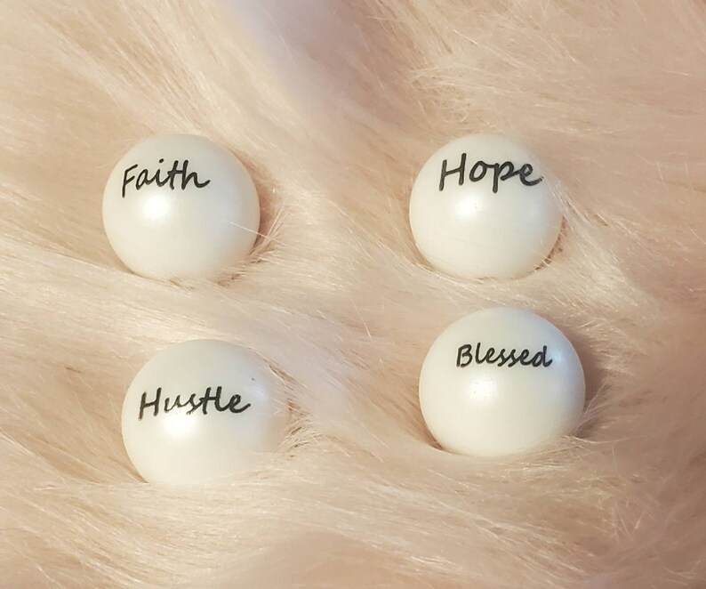 Puede incluir: Cuatro adornos redondos blancos con letras negras. Las palabras en los adornos son "Faith", "Hope", "Hustle" y "Blessed".
