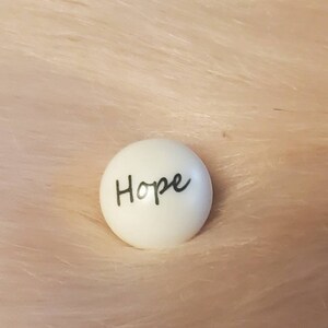 Puede incluir: Piedra redonda de cer&aacute;mica blanca con la palabra "Hope" escrita en cursiva negra.