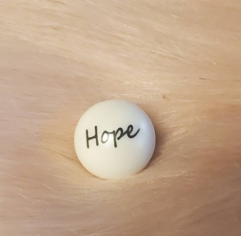 Puede incluir: Piedra redonda de cer&aacute;mica blanca con la palabra "Hope" escrita en cursiva negra.