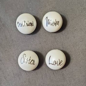 Puede incluir: Cuatro piedras ovaladas blancas con letras negras. Las piedras dicen "Best Friend", "Mom", "Sister" y "Love".