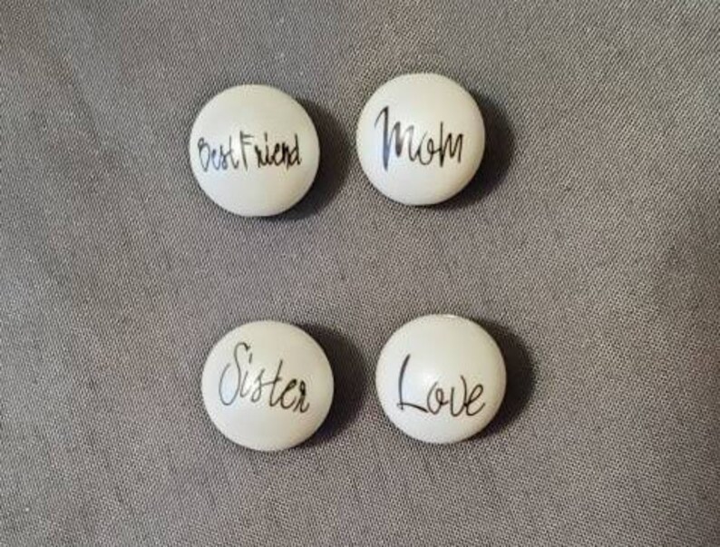 Puede incluir: Cuatro piedras ovaladas blancas con letras negras. Las piedras dicen "Best Friend", "Mom", "Sister" y "Love".