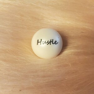 Puede incluir: Bola redonda blanca con la palabra "Hustle" escrita en cursiva negra.