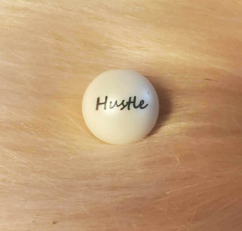 Puede incluir: Bola redonda blanca con la palabra "Hustle" escrita en cursiva negra.
