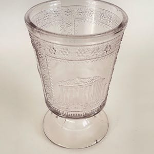 May include: A clear, pale pink glass goblet with a footed base. The goblet has a decorative pattern around the rim and a raised design of a building on the side.