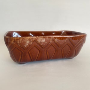 May include: A rectangular brown ceramic baking dish with a geometric diamond pattern. The dish has a glossy finish and is set against a white background. The dish is approximately 10 inches long.