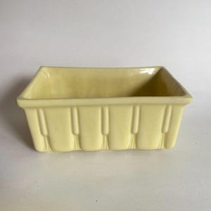May include: A rectangular, pale yellow ceramic planter. The planter has vertical ribbed detailing on the sides and a smooth, open top. The interior is a slightly darker shade of yellow.