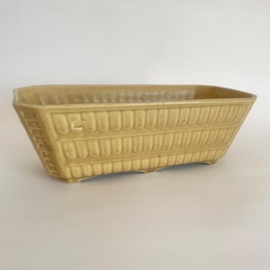 May include: A rectangular, light yellow ceramic planter with a textured, basket-weave design. The planter has a slightly curved base and small feet. The sides are angled, and the top edges are smooth. A classic piece for indoor or outdoor plants.