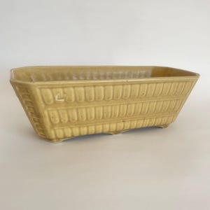 May include: A rectangular ceramic planter with a glossy mustard yellow glaze. The exterior features a repeating vertical ribbed pattern along the sides and ends, with a smooth rim and small feet supporting the base of the vessel.